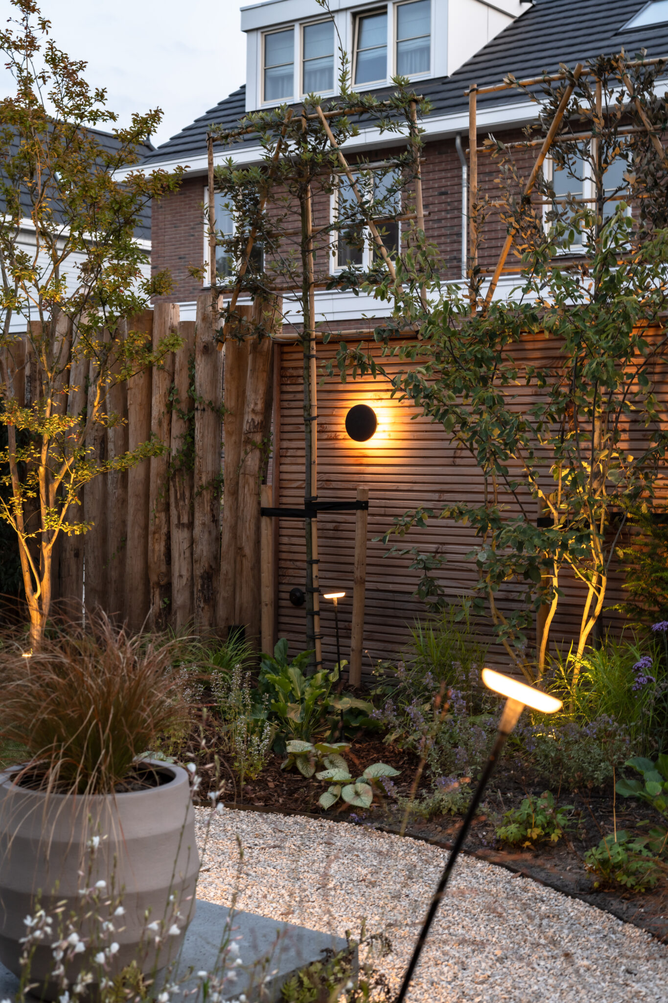 Avondtuin met een ronde wandlamp, verlichte bomen en een staande borderlamp.